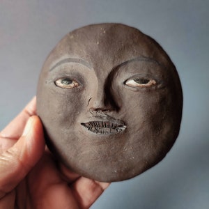 Könnte beinhalten: Eine dunkelbraune Keramikskulptur eines Gesichts mit bemalten Augen und leicht geöffnetem Mund.