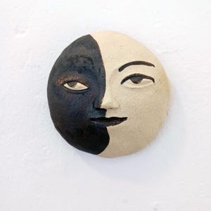 Könnte beinhalten: Eine kreisförmige Wandmaske aus Keramik mit einem geteilten Gesicht. Die linke Seite ist mit einer dunklen, texturierten schwarzen Glasur versehen, während die rechte Seite matt und cremeweiß ist. Das Gesicht zeigt geschnitzte Augen, eine Nase und einen schmalen Mund.
