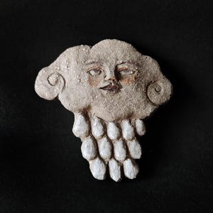 May include: A ceramic sculpture of a face with a cloud-like head and a white beaded necklace. The face has a neutral expression with closed eyes and a slightly parted mouth.