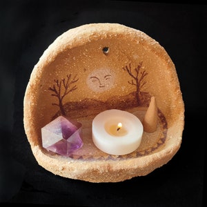Puede incluir: Un quemador de incienso de arcilla color canela con textura en forma de semicírculo, que presenta un paisaje pintado con una luna y árboles desnudos. Dentro hay un cristal morado facetado, una vela de té encendida y un cono de incienso marrón.