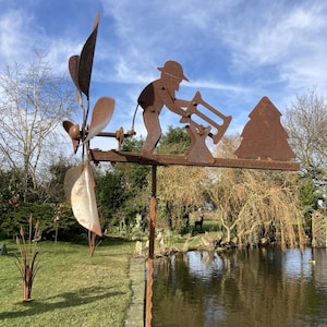 May include: A weathered metal garden ornament depicting a figure playing a trumpet, a tree, and a pinwheel. The sculpture is mounted on a tall, textured pole, set against a backdrop of a pond and a blue sky with some clouds.