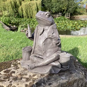 May include: A detailed statue of a frog in a plaid jacket, holding a bottle and a sandwich. The frog is seated on a large rock, with a small basket and a container nearby. The statue is grey and set outdoors.