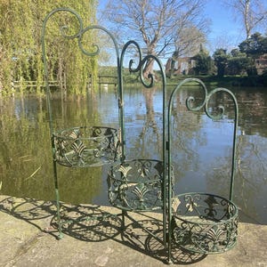 May include: A three-tiered, green metal plant stand with ornate scrollwork and basket-style holders. The stand is outdoors, set against a backdrop of a pond and trees. The plant stand is approximately 127 cm tall.