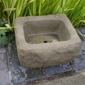 May include: A rectangular stone bird bath with a rough, textured surface. The bird bath is filled with water and is sitting on a patio.