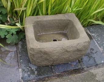 May include: A rectangular stone birdbath with a rough, textured surface. The birdbath is filled with water and is sitting on a patio.