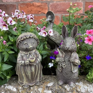 May include: Three garden figurines, a hedgehog, a rabbit, and a duck, are arranged on a stone surface. The hedgehog is holding a teacup, the rabbit is holding a carrot, and the duck is wearing a hat. The figurines are painted in a rustic, weathered style.