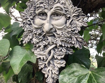 Op de afbeelding: Een gedetailleerde, gebroken witte Green Man gezichtssculptuur met een baard en blad details. De sculptuur is gemonteerd op een boomstam en omgeven door groene klimopbladeren. Het gezicht heeft een serene uitdrukking.