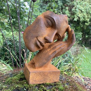 May include: A rust-coloured sculpture of a human face resting on a hand, mounted on a square base. The surface has a weathered, textured appearance with deep brown and orange tones, depicting a serene expression with closed eyes and a hand.