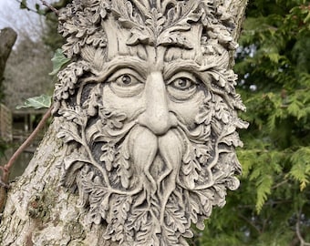 May include: A grey stone sculpture of a Green Man's face with a leafy beard and crown. The sculpture is mounted on a tree trunk.
