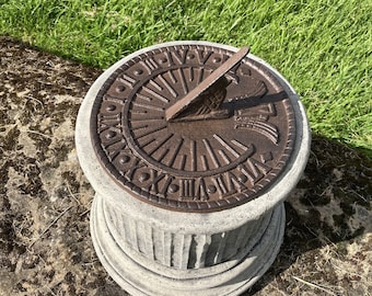 May include: A brown metal sundial with Roman numerals on a gray concrete base. The sundial is sitting on a bed of green grass.