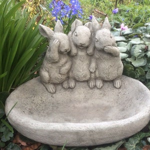 May include: Three grey stone rabbits with their eyes, ears and mouths covered, sitting on the edge of a grey stone oval bird bath.