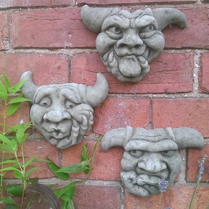 May include: Three grey stone gargoyle sculptures with exaggerated features, including large noses, wide mouths, and protruding tongues, mounted on a brick wall.
