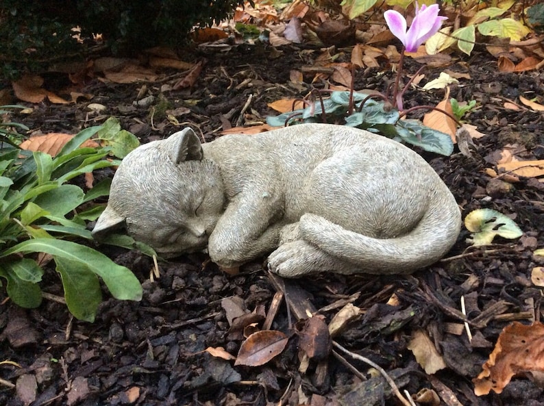 May include: A gray stone cat sculpture in a curled sleeping position. The cat's eyes are closed, and its paws are tucked in. The sculpture is surrounded by mulch, leaves, and green plants. A pink flower is visible in the background.