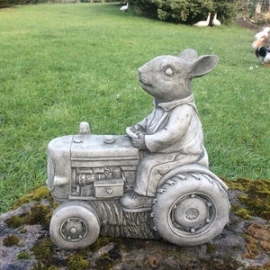 May include: A grey concrete garden statue of a rabbit driving a tractor. The rabbit is wearing a shirt and trousers. The tractor has a detailed engine and wheels.