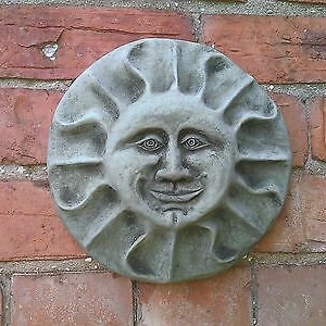 May include: A grey stone sculpture of a smiling sun face with a detailed design. The sun face is mounted on a brick wall.