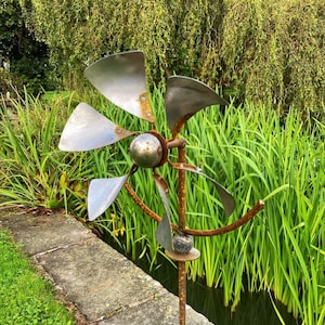 May include: A rusty metal garden sculpture in the shape of a flower with five blades. The sculpture is placed in a garden setting near a pond with green reeds.