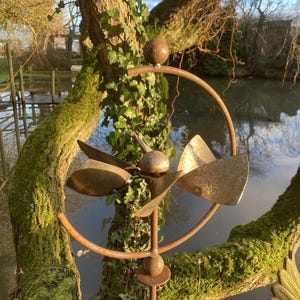 May include: A rusty metal garden sculpture with a spinning propeller design. The sculpture is mounted on a metal pole and is surrounded by green foliage.