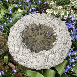 May include: A white concrete bird bath with a leafy design. The bath is filled with water and surrounded by blue flowers.