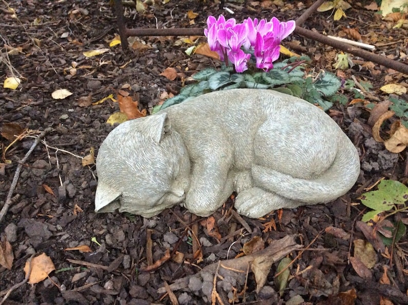 May include: A light gray, sleeping cat statue with detailed fur texture. The cat is curled up, with its head resting on its paws. The statue is placed outdoors, near pink flowers and fallen leaves.