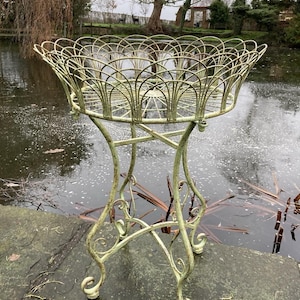 May include: A vintage, pale green metal plant stand with an ornate, openwork basket. The stand features a scrolled base and a crisscross design supporting the basket. The stand is positioned near a body of water.