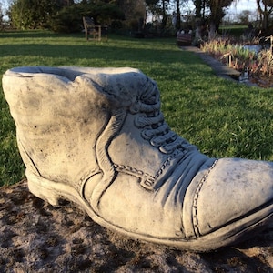 May include: A grey concrete boot planter with a lace-up design. The boot is sitting on a rock in a garden setting.