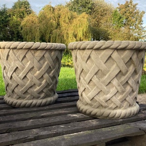 May include: Two large, grey concrete planters with a woven design. The planters are sitting on a wooden pallet.