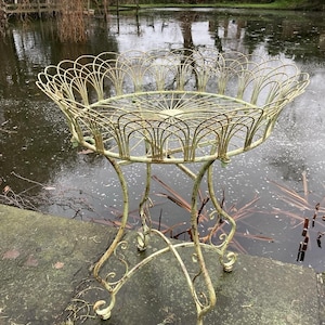 May include: A vintage, pale green metal plant stand with an ornate, openwork design. The stand has a circular top with a scalloped edge and curved legs with scrolled feet. It is positioned near a pond.