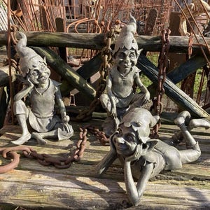 May include: Three grey stone garden gnomes with pointy hats and mischievous expressions. The gnomes are sitting on a wooden bench with rusty chains.