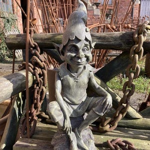 May include: A grey stone garden gnome statue with a pointed hat and a mischievous grin. The gnome is sitting on a rock with its legs crossed. The statue is surrounded by rusty chains and metal rods.