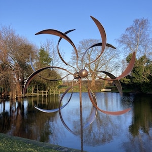 May include: A large metal wind spinner with multiple curved blades in a rusty brown colour. The spinner is standing in a grassy area next to a pond with a blue surface.