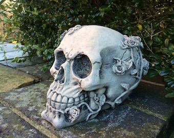 May include: A white concrete skull with roses sculpted into the bone. The skull is sitting on a brick surface.