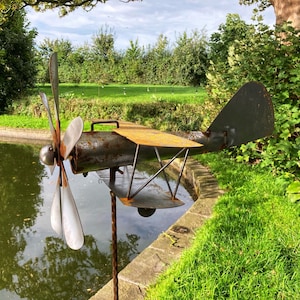 May include: A rusty metal airplane wind spinner with a propeller and wings. The spinner is mounted on a metal pole and is placed in a grassy area near a pond.