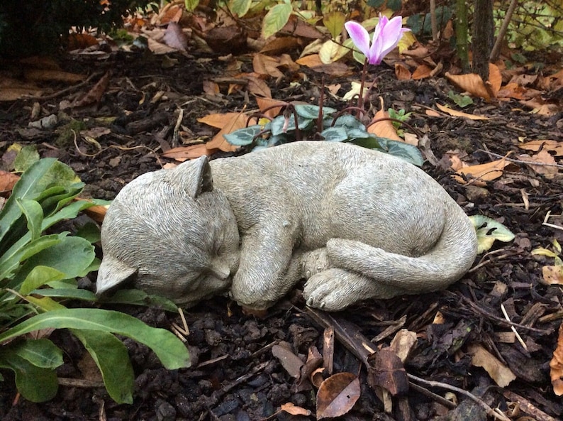 May include: A gray stone cat sculpture in a curled sleeping position. The cat is lying on a bed of mulch and fallen leaves, with green plants and a pink flower in the background. The sculpture has detailed features.