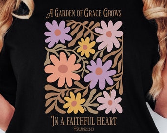 Puede incluir: Camiseta negra con un diseño floral y el texto "A GARDEN OF GRACE GROWS IN A FAITHFUL HEART" y "PSALM 92:12-13". El diseño incluye flores melocotón, moradas y amarillas. La camiseta también tiene el texto "Loop's Crafty Creations".