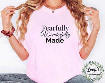 May include: A pale pink t-shirt featuring the words "Fearfully Wonderfully Made" in black script and bold lettering. The top has a crew neck design.