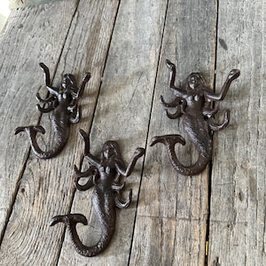 May include: Three cast iron mermaid wall hooks in a dark brown finish. Each hook features a mermaid with outstretched arms and a fishtail. The hooks are mounted on a weathered wooden surface, adding a rustic touch to the decor.