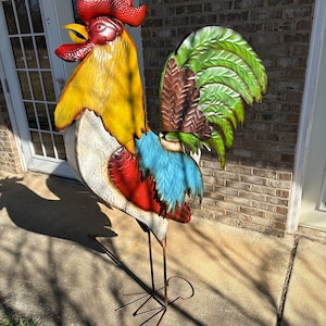 May include: A colorful metal rooster sculpture with a red comb, yellow head, white body, blue and red tail feathers, and green and brown wing feathers. The rooster is standing on a metal stand with four legs.