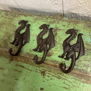 May include: Three cast iron dragon hooks in a dark brown finish. Each hook features a dragon with outstretched wings and a curved tail. The hooks are mounted on a distressed green wooden surface.