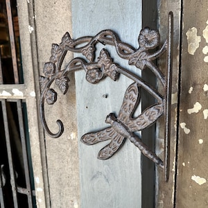May include: A cast iron plant hanger with a dragonfly design and a vine motif. The hanger is painted in a dark brown color and has a hook for hanging.