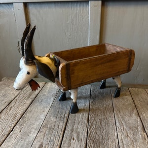May include: A white and black painted metal goat sculpture with a wooden box on its back. The goat has a red tongue sticking out and black horns. The wooden box has a rustic brown finish.
