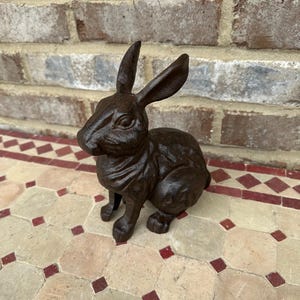 May include: A detailed, dark brown cast iron rabbit statue. The rabbit is in a seated position with its ears perked up. The statue is on a patterned tile surface with a brick background.