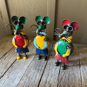 May include: Three metal mice figurines with colorful paint. Each mouse has a different color scheme, including red, yellow, green, and blue. The mice are standing on a wooden surface.