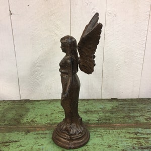 May include: A cast iron angel statue with wings, standing on a pedestal. The angel is brown and has a distressed finish.