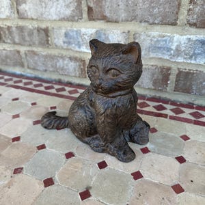 May include: A brown cast iron cat doorstop. The cat is sitting with its tail curled around its legs. The doorstop is on a tiled floor.