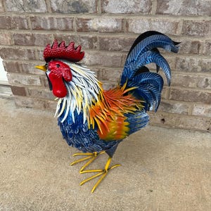 May include: A colorful metal rooster sculpture. The rooster features a red comb and wattle, white and blue feathers, and yellow legs. The sculpture is painted with vibrant shades of blue, orange, and yellow.