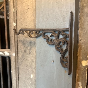 May include: Ornate, dark brown cast iron plant hanger. The bracket features a decorative scroll design and is mounted on a light gray wall. The hanger extends outward with a curved end for holding a pot or basket.