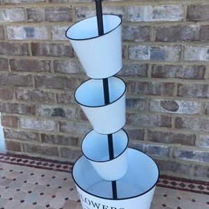 May include: White metal tiered planter with black metal stand and a black rim around each planter. The bottom planter has the text "FLOWERS & GARDEN" printed on the side.