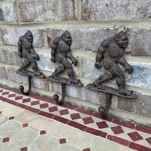 May include: Three cast iron Bigfoot hooks mounted on a brick wall. Each hook features a detailed Bigfoot sculpture with a hook extending from the bottom.