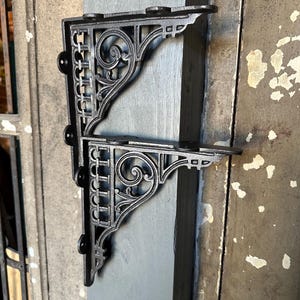 May include: Two black, ornate cast iron shelf brackets. Each bracket features a decorative scroll design and a series of small, round accents. The brackets are mounted on a gray and brown surface, suggesting a vintage or antique aesthetic.