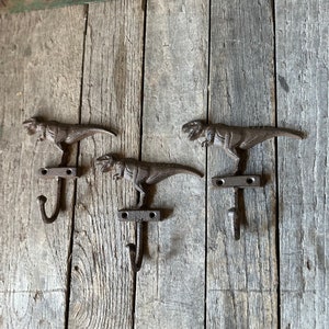 May include: Three cast iron dinosaur hooks mounted on a weathered wooden wall. Each hook features a dinosaur with an open mouth and a curled tail. The hooks are a dark brown color and have a rustic finish.